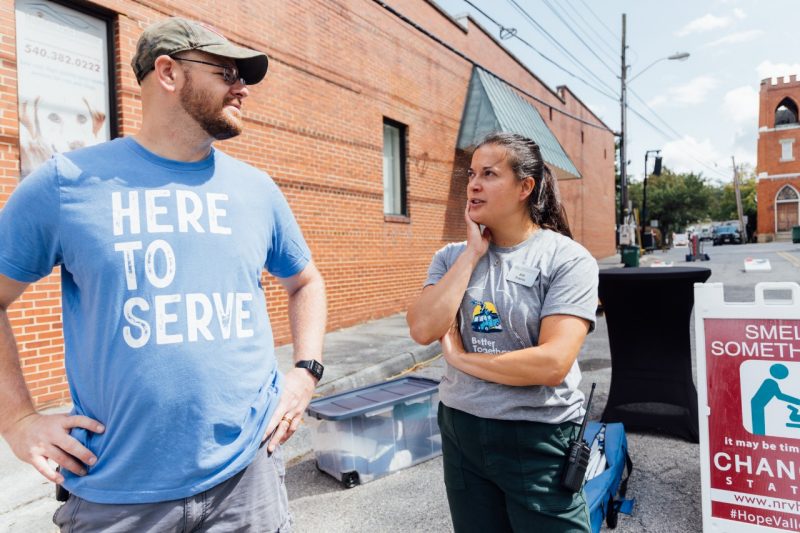 A man in a blue t-shirt that says "Here to Serve" speaks with a woman in a gray t-shirt outside a brick building.
