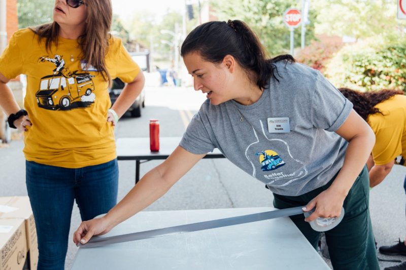 Elli marks out a folding table with duct tape. Two volunteers in yellow food truck rodeo shirts work in the background.
