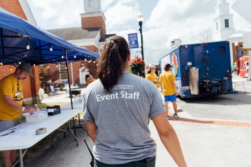 Elli walks up the sidewalk between tents and food trucks, her back is to the camera, the back of her shirt has the DCI logo on it and it says "EVENT STAFF".