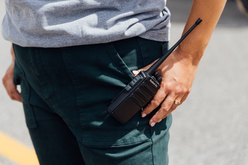 A close up of a walkie-talkie on the outside of a pants pocket, a hand rests on it.