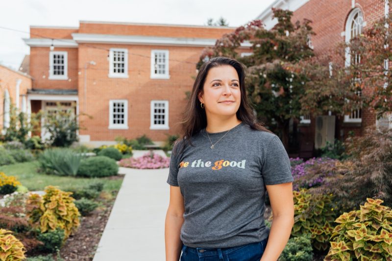 A woman in a gray t-shirt poses in front of a beautiful garden filled with hostas, coleus, and other native plants.