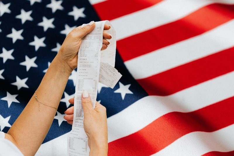 A shopper holding a grocery receipt, American flag in background. Images courtesy Pexels.