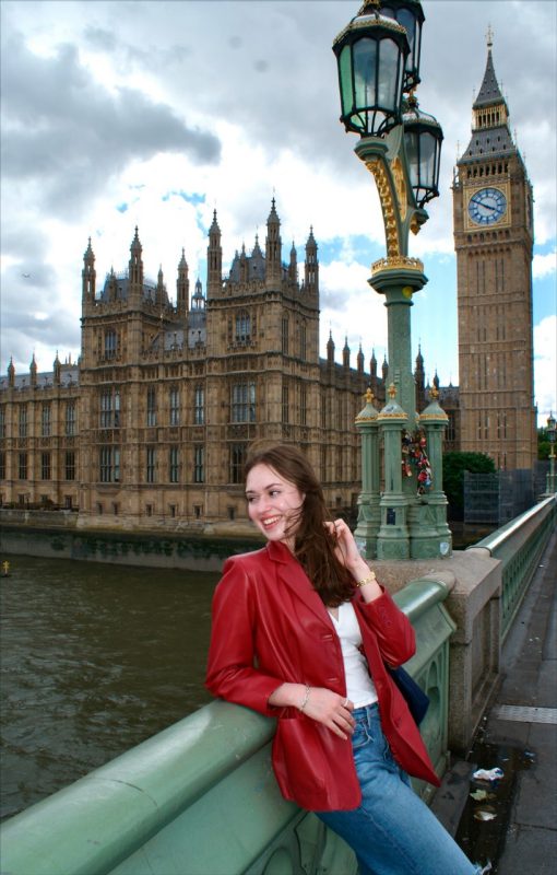 Carol Rector London Calling A woman with long bdrown hair and a red jacket stands on a bridge with Big Ben in the background.