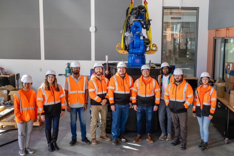 (From Left) Anasuya Kamakshi Tippabhotla, Kereshmeh Afsari, Sathya Narayanan Vaaradharaj, Mike White, Alex Brand, Andrew McCoy, Akhileswar Yanamala, Nathan King, and Vida Babajaniniashirvani stand with new 3D printer inside Hitt Hall's Innovation Lab. Photo by Ashley Williamson for Virginia Tech. A group of people in reflective orange shirts and hardhats stand in front of large 3D printer with robotic arm.