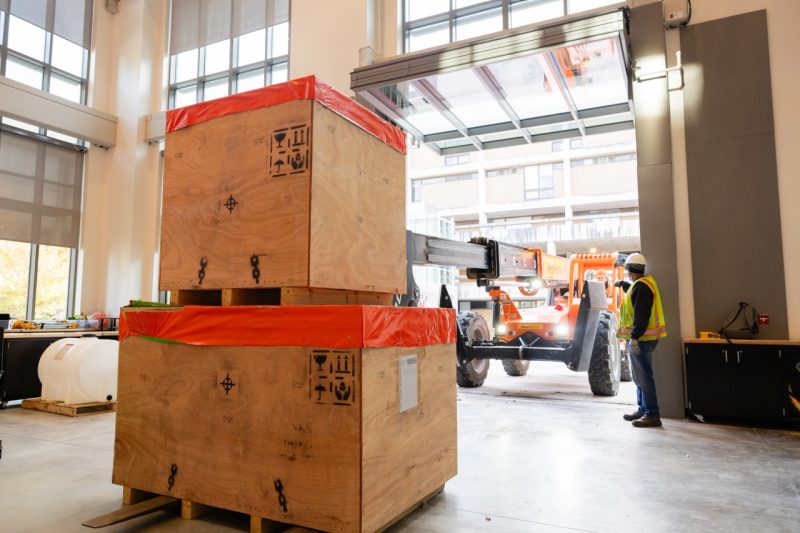 Boxes containing the 3D printer's parts arrive into Hitt Hall's Procon Innovation Center to be assembled. Photo by Ashley Williamson for Virginia Tech. Large wooden boxes sit on the left while a crane brings them into the lab.