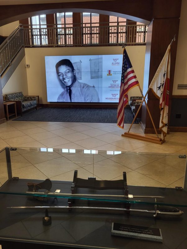 Lindsay Cherry '58 donates saber and ring to the Black Cultural Center A Corps of Cadets saber and ring on display in a glass case in front of a large screen depicting a historical photo of Lindsay Cherry '58.