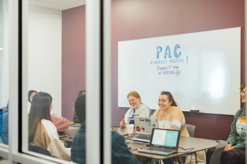 Eight or nine college students gather around a table with laptops open; the words "PAC Biweekly Meeting" are written on the white board in the background.