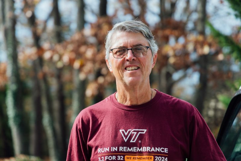 A portrait of John Galbraith, smiling at the camera and wearing a maroon 'Run in Remembrance' shirt, stands outside surrounded by trees with autumn leaves.