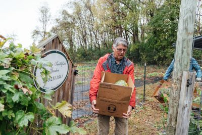 John Galbraith, wearing a red jacket and damp with the cold rain, carries a cardboard box filled with squash near a wooden garden sign reading 'Historic Smithfield Garden.' Another volunteer works in the background.