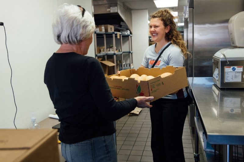 Marilyn Galbraith and Izzy Largen in a food pantry pass a cardboard box filled with winter squash, smiling at each other. Shelves with supplies and kitchen equipment line the background.