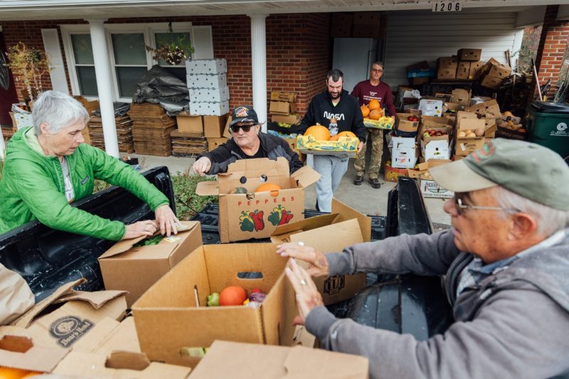Several volunteers load freshly gleaned produce into the back of a white pickup truck parked in the driveway of John Galbraith's house. The driveway is surrounded by stacks of boxes filled with fruits and vegetables, and the house serves as a storage site for the donations.