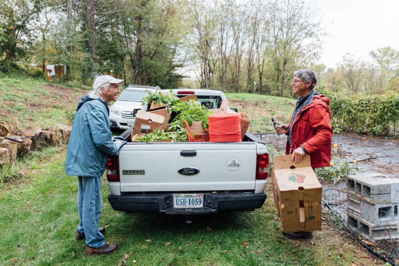 Master Gardener David McEwen and John Galbraith stand at the back of a pickup truck filled with boxes of fresh produce. John smiles while holding a box, and trees and greenery surround the scene. They are both wearing raincoats damp with cold rain.