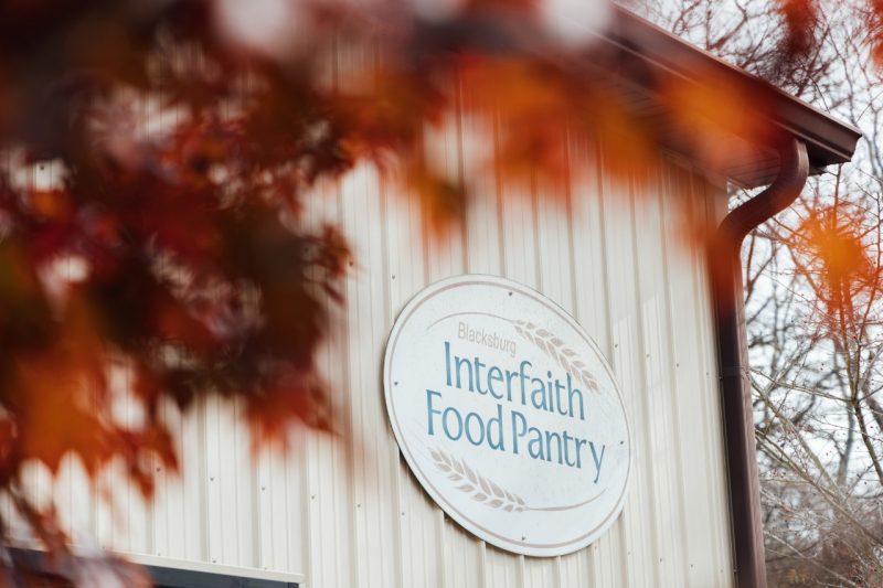 The oval sign for the Blacksburg Interfaith Food Pantry is visible on a beige metal building, framed by vibrant red autumn leaves in the foreground.