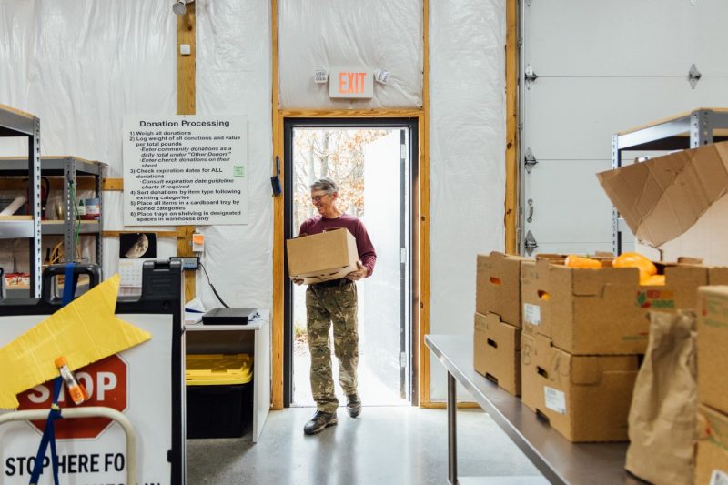 John Galbraith carries a box of gleaned produce into the Interfaith Food Pantry through an open door. Boxes of produce are stacked on a nearby table.
