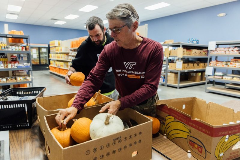 John Galbraith, wearing a maroon shirt, and his son, in a black sweatshirt, sort small pumpkins and squashes into boxes at the Interfaith Food Pantry in Blacksburg, surrounded by shelves stocked with food supplies.