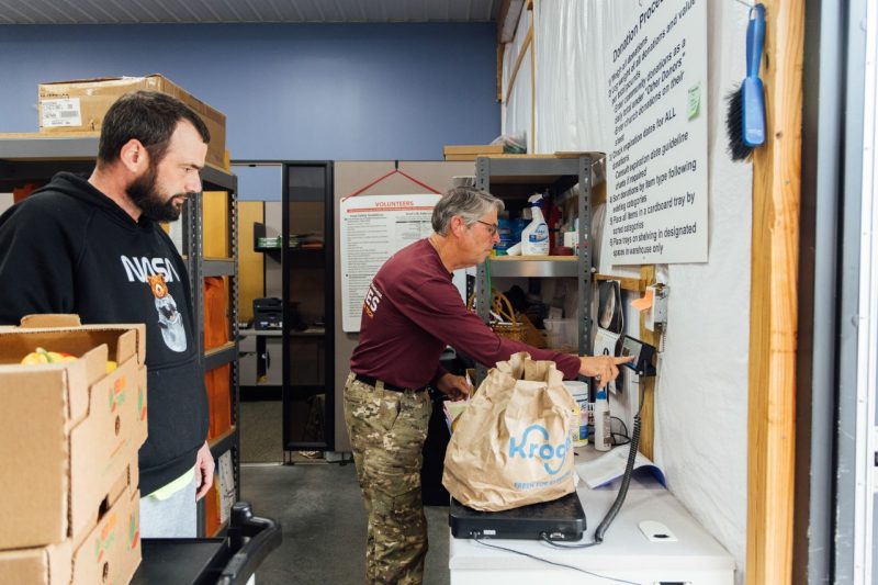 John Galbraith weighs a bag of fresh produce on a scale at the Interfaith Food Pantry while his son observes nearby. Donation guidelines are posted on the wall in the background.