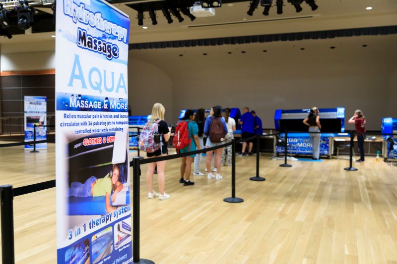 A line of students waits in the background for a row of blue-colored aqua massage tables. A sign in the foreground depicts a student with headphones lying in one of the tables and advertises a "Hydrotherapy Massage" service that can "Relieve muscular pain & tension and increases circulation with 36 pulsating jets to temperature controlled water while remaining clothed & dry."