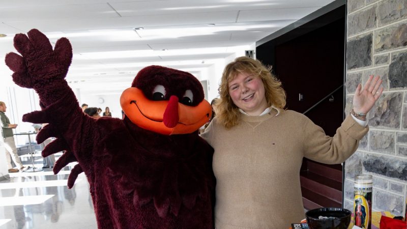 Kate Stanko and HokieBird The HokieBird mascot and a blond woman in a beige sweater wave viewers.