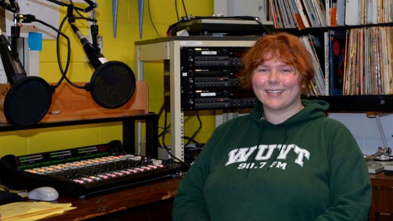 Kate Stanko WUVT A red-haired woman wearing a green WU'VT 90.7 FM sweat shirt sits behind a microphone. Shelves filled with record albums are behind her.
