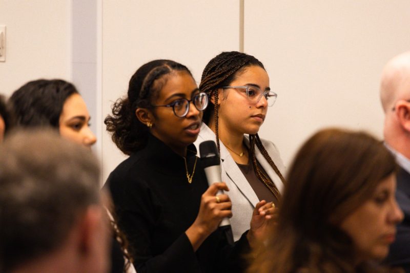 Men and women listen to a panel discussion. A female attendee speaks into the microphone to ask a question.