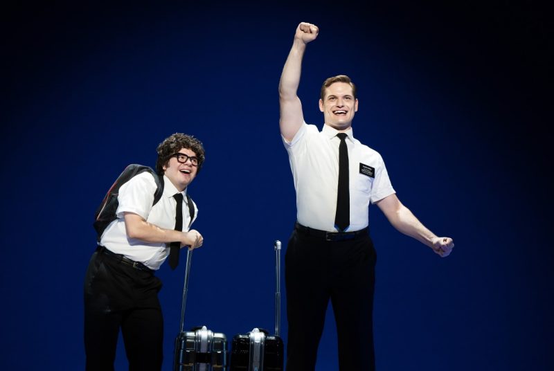 In a scene from the Broadway touring production of "The Book of Mormon," two men dressed as missionaries stand against a solid blue background, wearing white short-sleeve shirts, black ties, and black trousers. One man, on the right, raises his fist triumphantly and smiles confidently, while the other, on the left, leans slightly forward, wearing glasses, a backpack, and holding the handle of a rolling suitcase, looking enthusiastic and amused. Both have a playful and animated demeanor, suggesting a theatrical or comedic setting.