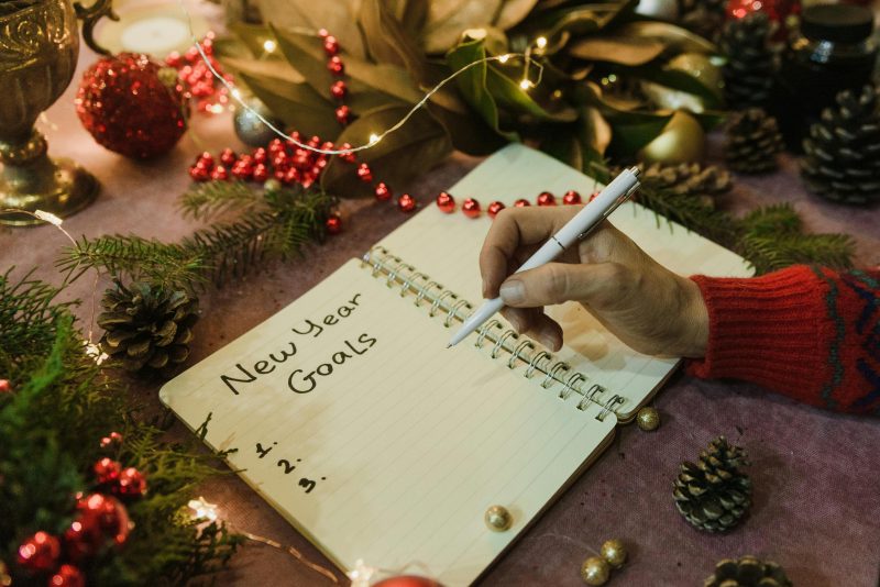 A hand writing in a notebook that says "New Year Goals," with holiday decor in the background.