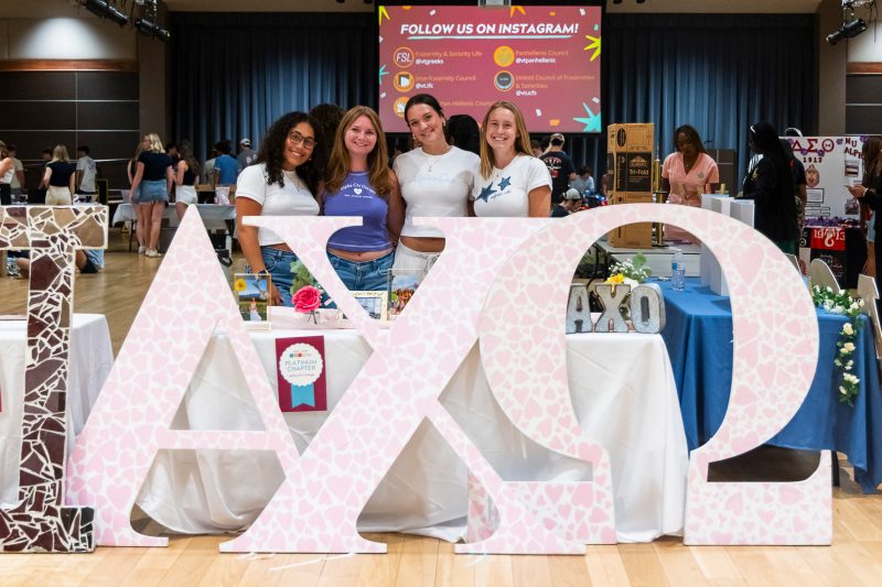 Spring Showcase Four women in t-shirts advertising their sorority, Alpha Chi Omega, stand behind a table and a large wooden sign with their Greek letters. Behind them, you can see other tables in a large space where students are chatting.