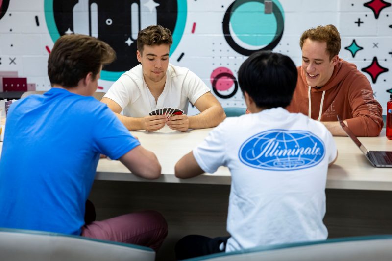Breakzone Uno Four students in casual dress, two with their backs turned and two facing the camera, play a game of Uno at a light brown table. The wall behind them has a colorful mural of pink and blue overlapping shapes and a large illustration of black headphones.