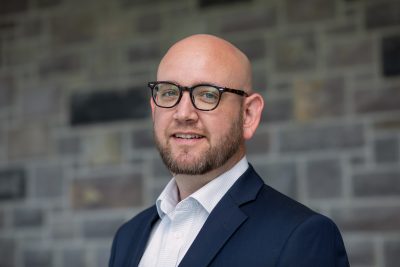 Portrait of Travis Jessee wearing a blue jacket and a white button down shirt smiling.