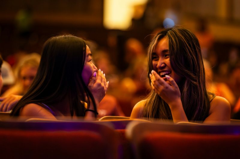 Two students are dimly lit in orange light, laughing with each other while partially covering their mouths with their hands. A crowd of students is seated behind them, and the tops of empty red-cushioned seats are in the foreground.