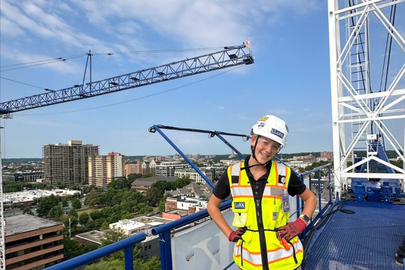 A woman wearing protective gear and a yellow vest stands atop construction scaffolding.