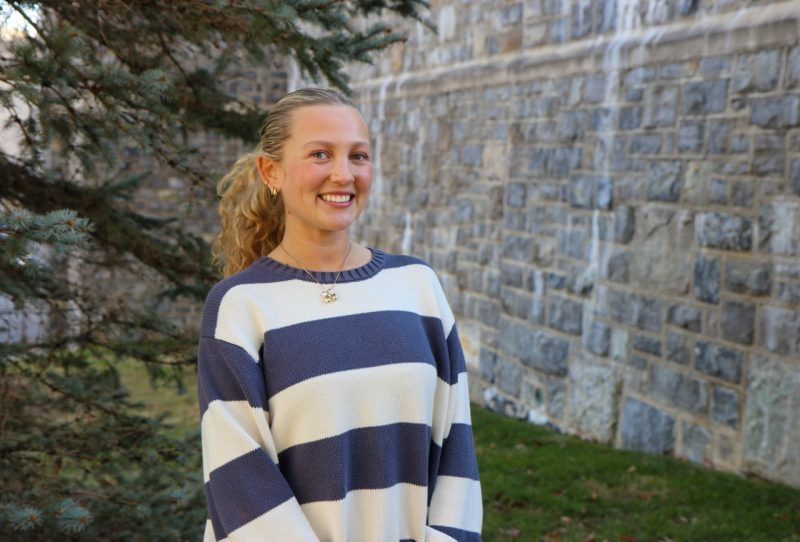 A young woman with blonde hair in a pony tail wears a blue and white striped sweater. She is standing outside of a building made of Hokie Stone, and in front of a pine tree.