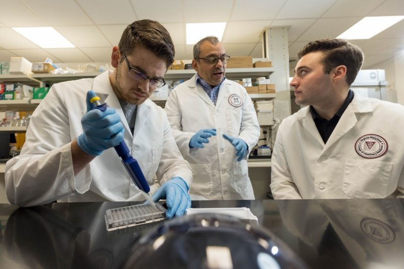Ramirez Zoetis Roger Ramirez-Barrios (at center) speaking with students (at right and left) while they do laboratory work. The student at left is holding a pipette.