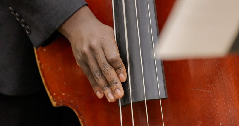 VT Jazz Ensemble A macro shot of a slightly weathered stand-up bass with a red wood finish and a dark grey fingerboard. A students' hand with a charcoal grey suit cuff rests on the left-most string.