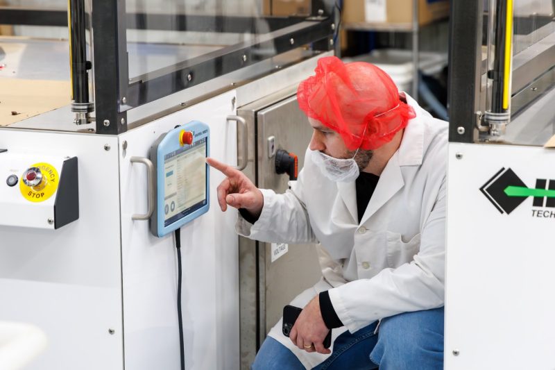 A man in hair net crouches to program a machine.