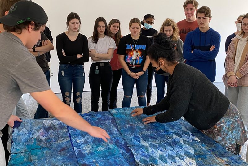 A group of students stands in a semi-circle around artist Yanira Collado who is demonstrating or assembling a large, textured artwork made of layered blue panels with geometric patterns. The artist, dressed in black, is carefully arranging the pieces, while a student in a cap reaches out to assist. The students observe attentively, some with arms crossed, in a Moss Arts Center gallery space..