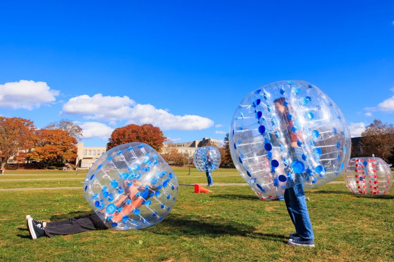 Bubble Soccer There are three students wearing body-sized inflatable bubbles, two in the foreground and one in the background. One of the foreground students is laying down with their hands on their heads. An unoccupied bubble sits on the right side of the image. There are orange cones on the grass, and much of the background is blue sky with very little cloud cover in it. A few autumn trees and academic buildings are visible in the distance.