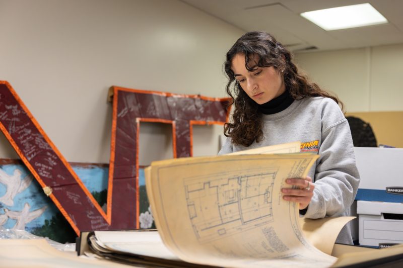 A young woman with long curly hair stands at a table in a library archive room, intently examining a large architectural drawing on yellowed paper. She is wearing a light gray sweatshirt and is surrounded by stacks of archival documents and storage boxes. Behind her, a large maroon and orange VT sign leans against the wall. The setting appears to be an organized workspace within Special Collections and University Archives at Virginia Tech.