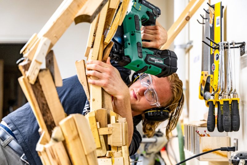 Ben Hornyak Using a Nail Gun A student wearing safety goggles and ear protection uses a nail gun to assemble a wooden animatronic sculpture in a workshop, surrounded by hand tools hanging on the wall.