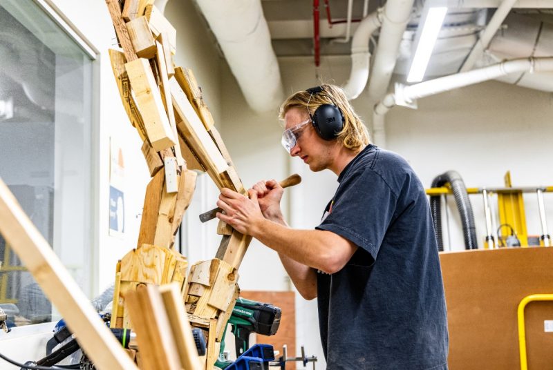 Ben Hornyak Using a File A student wearing safety goggles and ear protection uses a file to refine a large wooden animatronic structure in a workshop equipped with industrial tools and ventilation pipes.