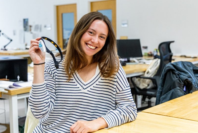 Megan Wysocki with Headset A student sits at a desk in a design studio, holding up a headset used for acoustic or cognitive research.