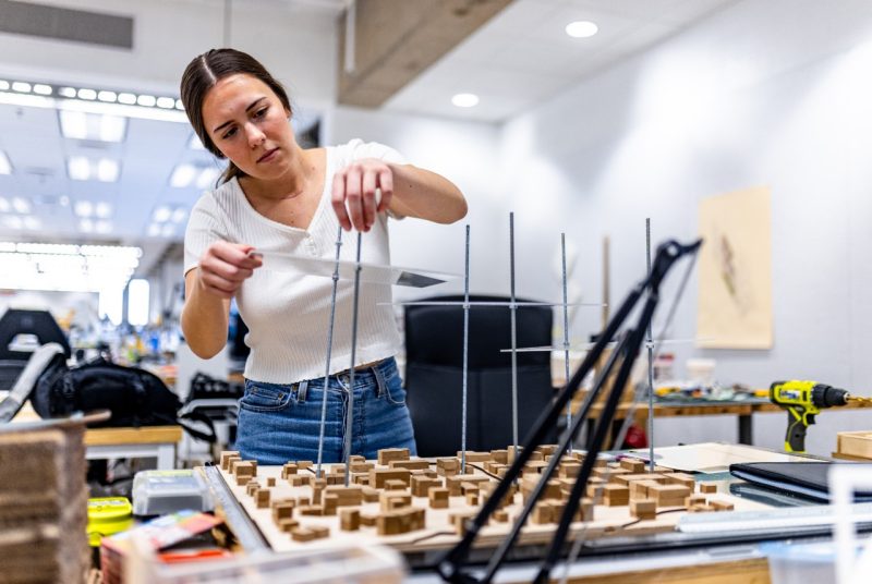 Olivia Kastor in the Studio A student adjusts thin rods on a detailed architectural model of a small town, working in a studio filled with tools, materials, and design equipment.