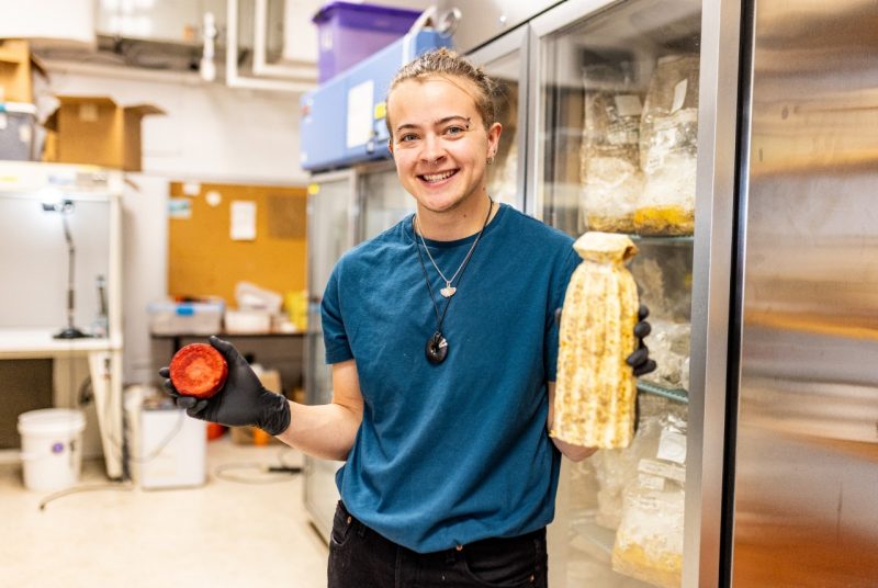 Avery Gendell in the Lab A student wearing latex gloves holds a round mycelium sample in one hand and a larger sculpted mycelium composite in the other, standing in a lab with refrigerated storage behind them.
