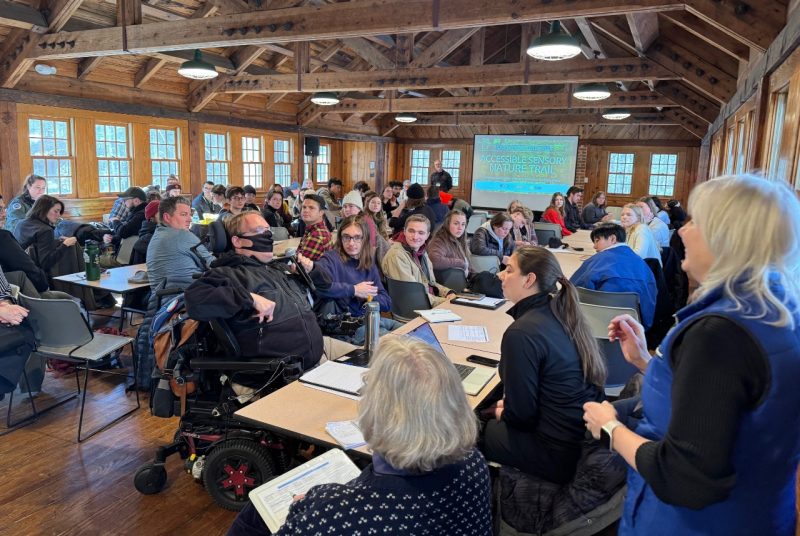 Students Meet with Community Members In a large room with wooden beams across the ceiling, many windows, and a projection screen at the front, a large group of students and community members sit at long tables.