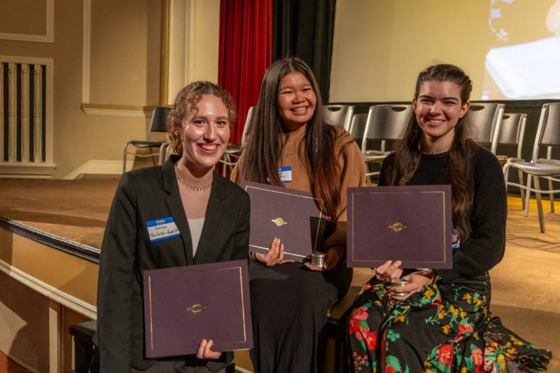 Three young women stand in front of a stage holding certificates, smiling.