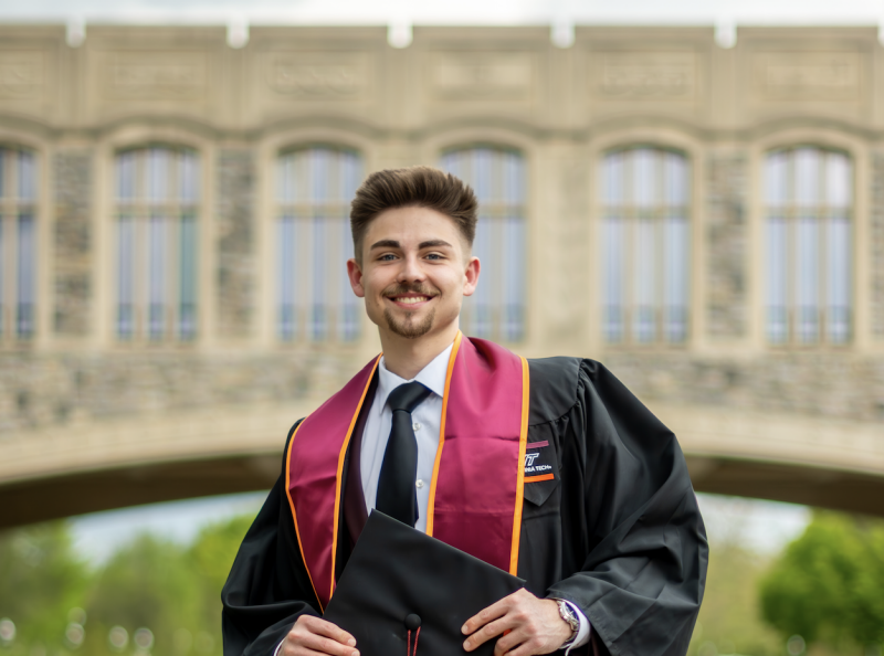 Giovanni Heater Grad Profile Photo of Giovanni Heater holding graduation cap in front of Torgersen Bridge