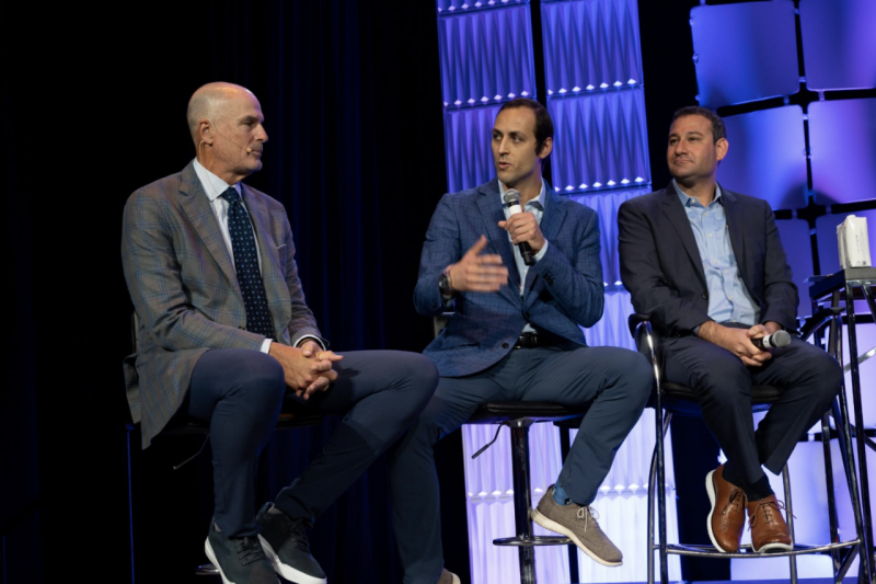 Three men in suits are seated in chairs on a stage. One is speaking into a microphone.