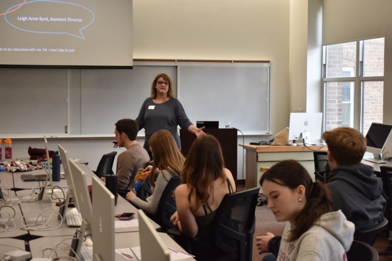 A row of students are seated at computers. An female instructor is at the front of the room, speaking. Above her is a large screen.