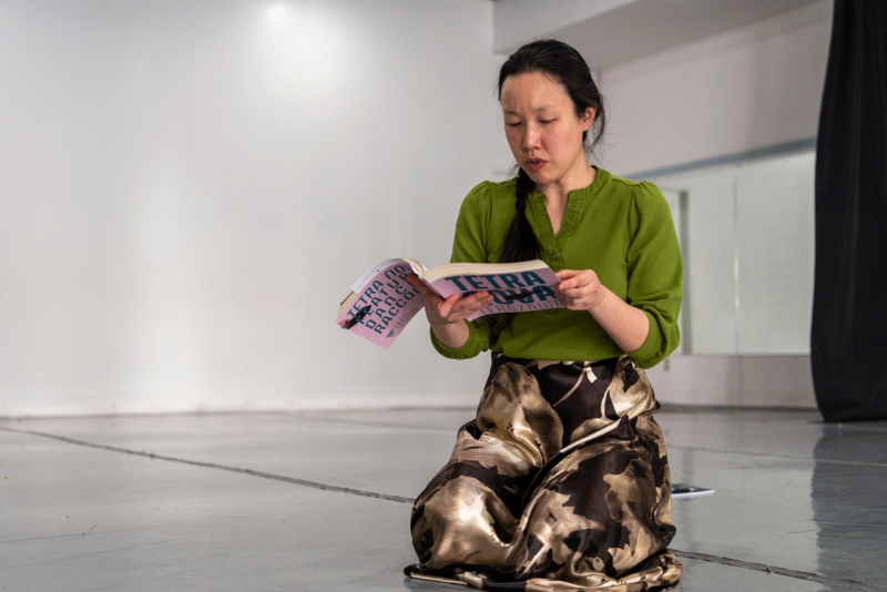 Sophia Terazawa A woman kneels on the floor while reading her book, 'Tetra Nova.' She wears a green shirt and brown flowered shirt.