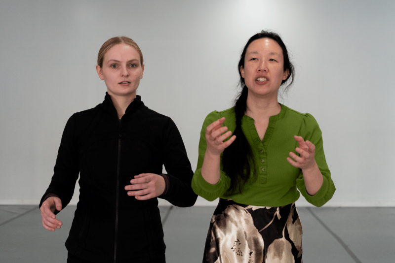 A woman in a black jacket and hair in a bun stands beside a woman with her hair braided.